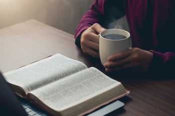 Hands holding a cup of coffee and a bible placed on a laptop