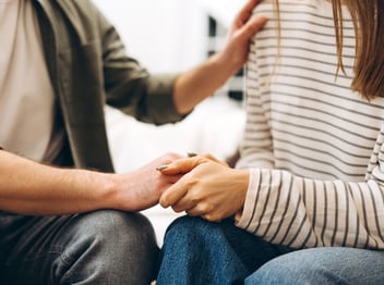 Two people sitting close together; one is placing a comforting hand on the other's shoulder and hands, suggesting support or reassurance.