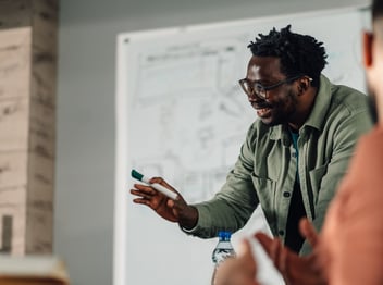 A man in glasses stands in front of a whiteboard, holding a marker and gesturing while speaking to someone out of frame.