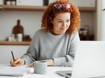 A woman with curly red hair writes in a notebook while looking at a laptop on a white desk, with a mug and earphones beside her.