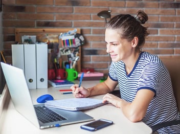 Person sitting at a desk, taking notes in a notebook while looking at a laptop, with office supplies and a phone nearby.