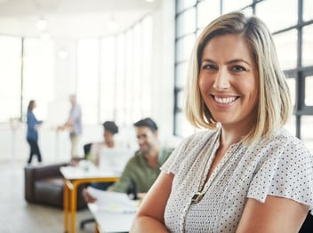 A woman with blonde hair smiles at the camera in a bright modern office, with three people working in the background.