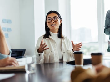 A woman wearing glasses speaks and gestures with her hands during a meeting at a table with coffee cups and documents in a bright office setting.